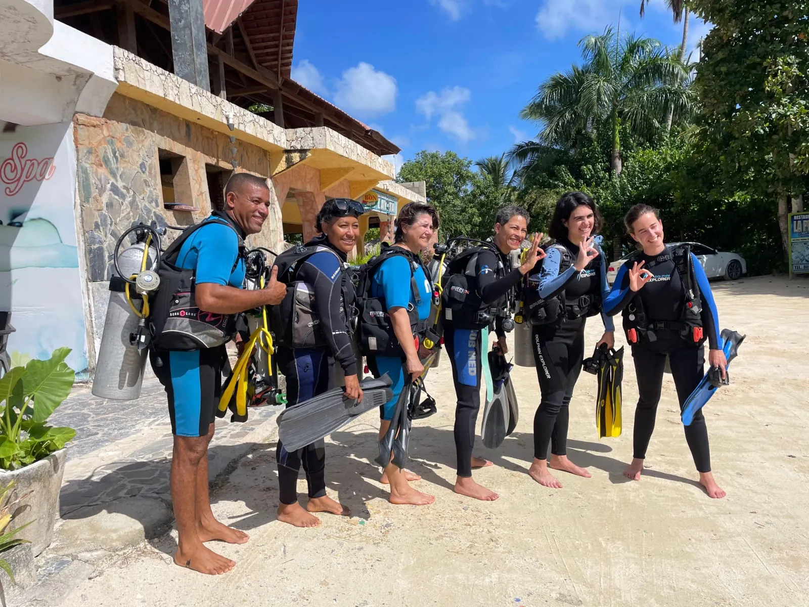 Six divers in wetsuits with tanks and fins, smiling and giving the OK sign on the sand