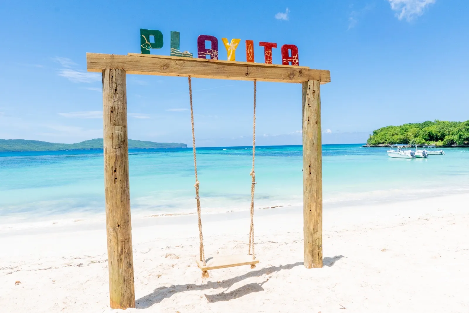 Swing with the PLAYITA sign on the white sand of Playa Playita, boats in the turquoise bay